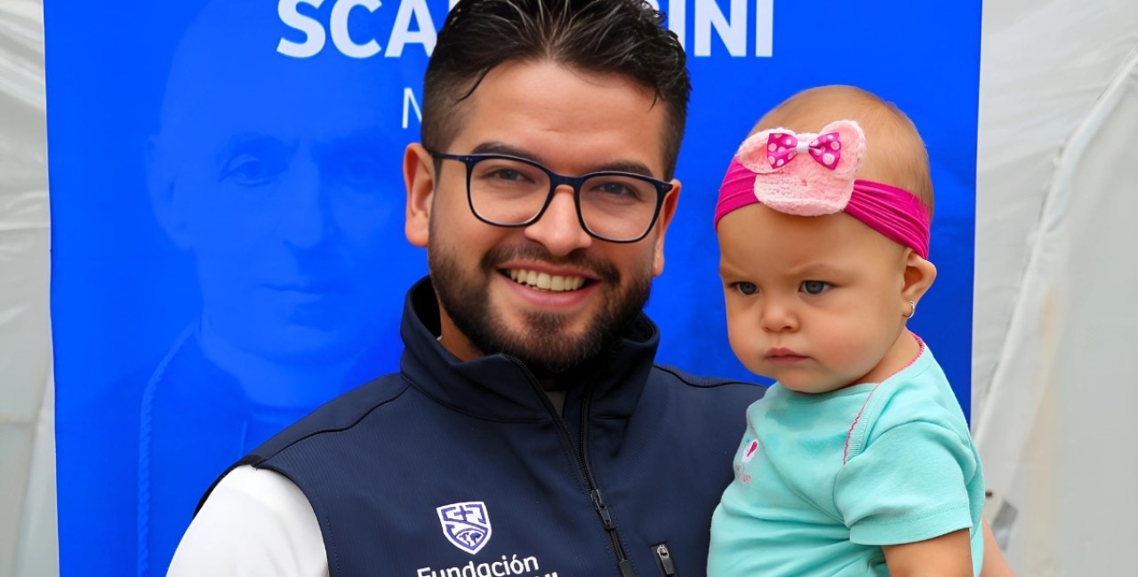 Un colaborador de la Fundación Scalabrini carga en brazos a una bebé frente a un cartel con la imagen del santo