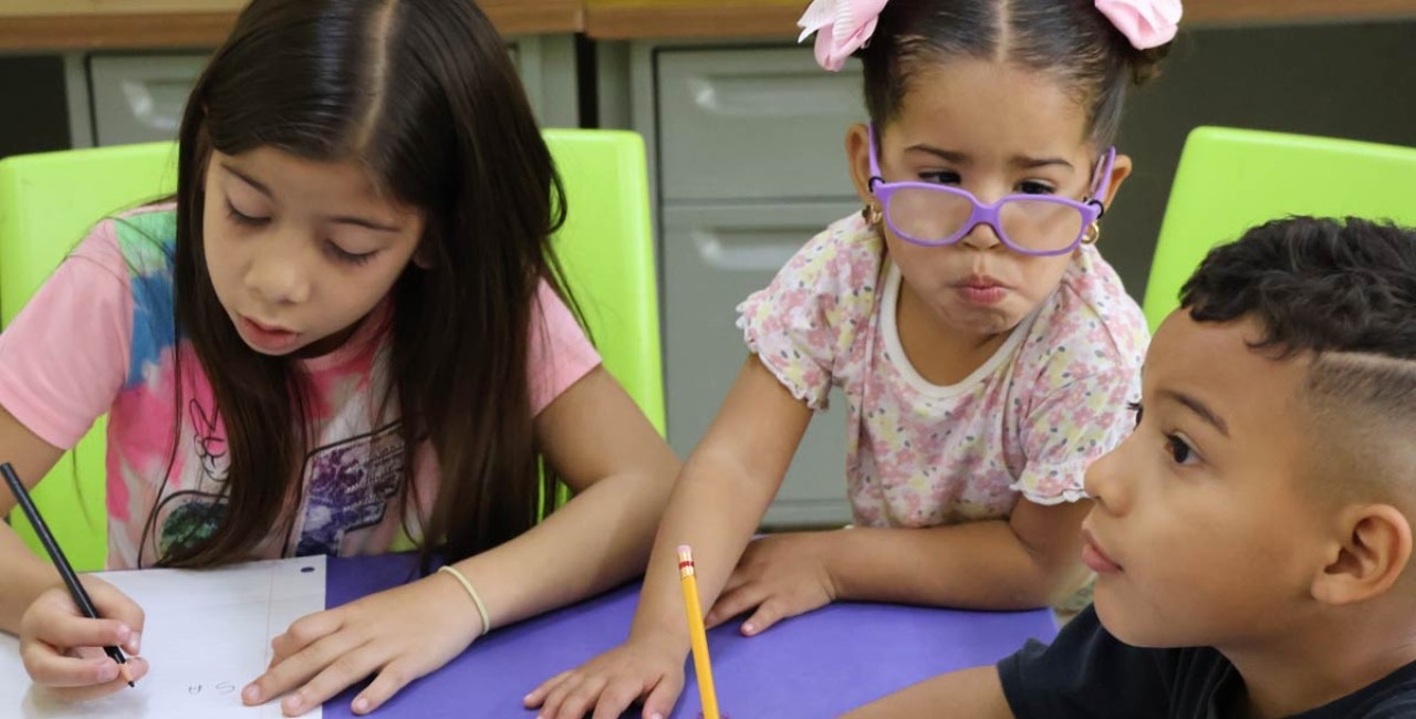 Tres niñas y niños trabajan juntos con lápiz y cuaderno en una mesa del aula de actividades